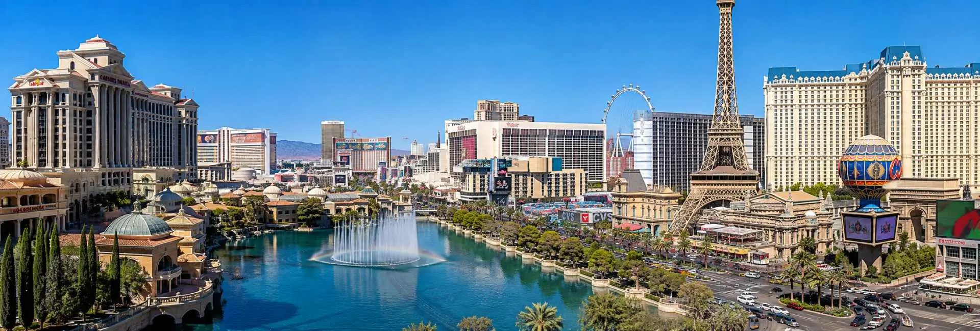 Las Vegas Strip with iconic resorts, Eiffel Tower replica, fountain and mountains under a clear blue sky